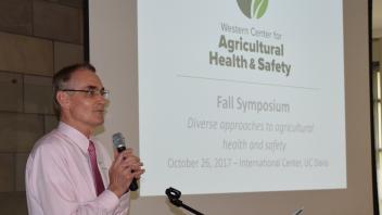 Dr Kent Pinkerton stands at podium with microphone screen in background shows slide with WCAHS logo