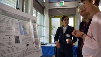three women stand smiling in front of a poster about collaboration mapping 