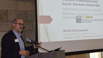 Dr Fadi Fathallah speaking at podium with slide in background showing presentation on robotic strawberry harvest aid research