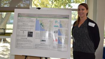 smiling woman with hands clasped behind back stands next to her research poster