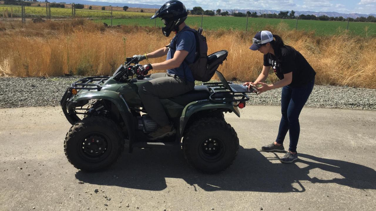 Person sitting on a green ATV with a researcher checking readings
